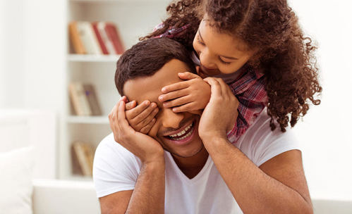 Daughter leaning over her dads back and keeping his eyes closed